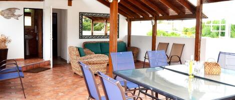Un patio techado con una mesa de vidrio, muebles de mimbre y vistas a la vegetación.