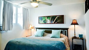Une chambre à coucher comprenant un lit, un ventilateur de plafond, une fenêtre avec des rideaux, un tableau accroché au mur et deux lampes de chevet.