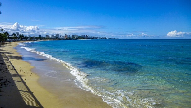 Une plage à l’eau d’un bleu limpide, une rive sablonneuse et, au loin, le profil d’une ville.