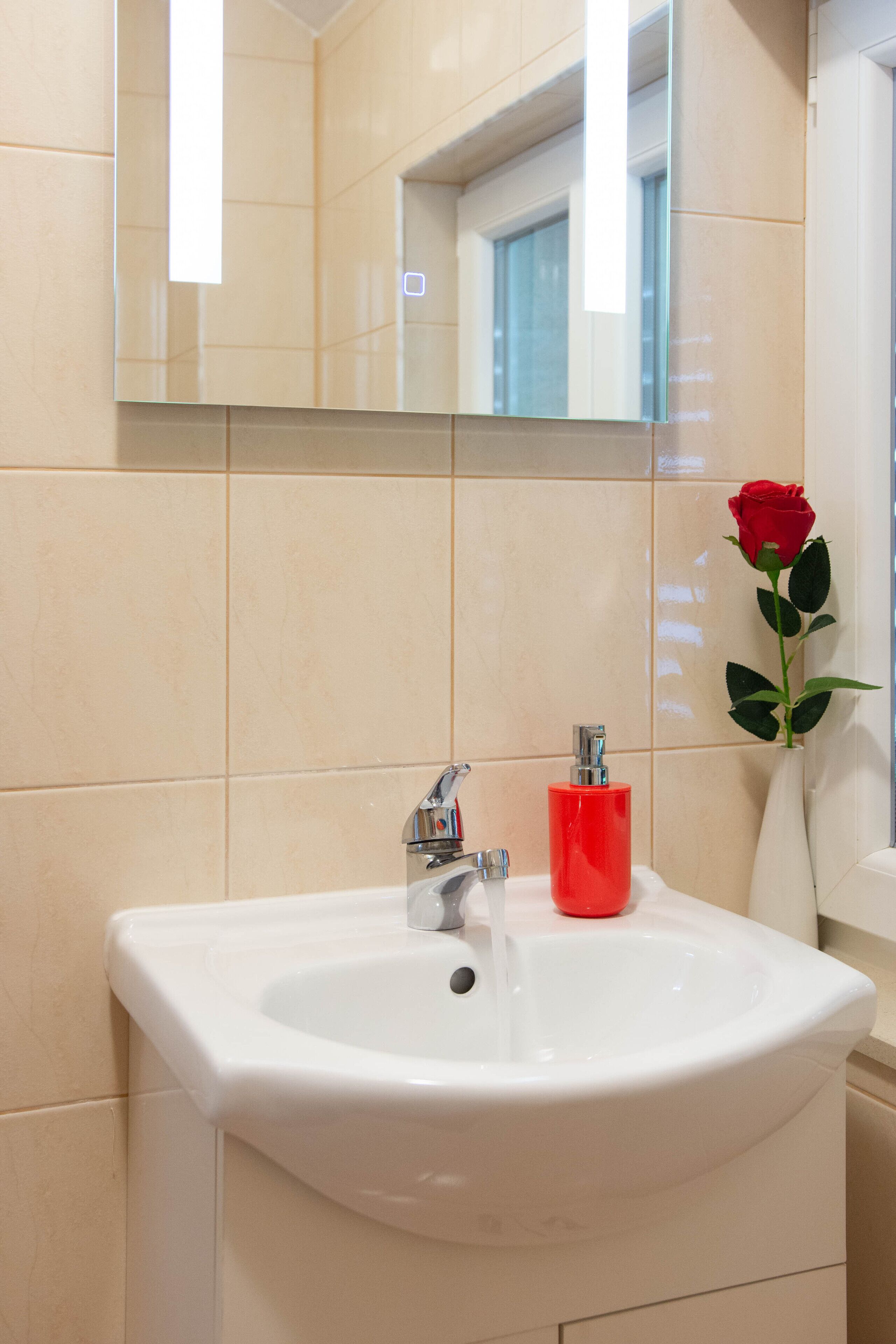 Un bagno con un lavandino bianco, un erogatore di sapone rosso e un vaso con una singola rosa rossa.