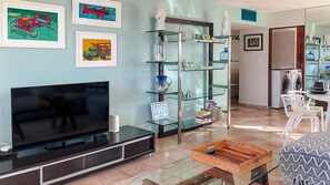 A living room with a flat-screen TV, a glass shelf unit, and a sign reading 'BACK TO THE BEACH'.