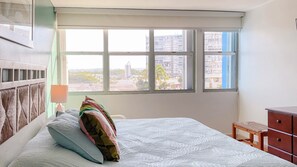 A bedroom with a bed, a nightstand, a dresser, and a window with a city view.