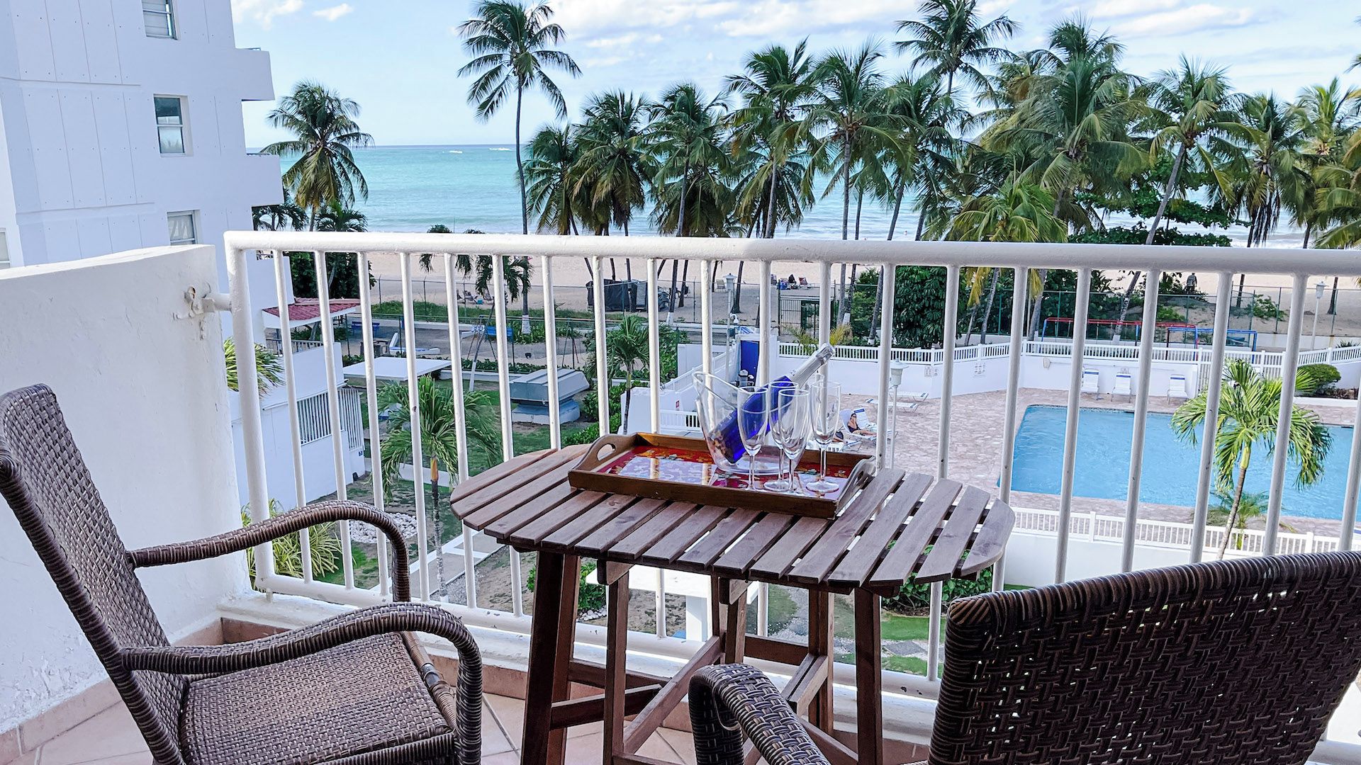 A balcony with wicker furniture, a table with a tray, and a view of a pool and palm trees.
