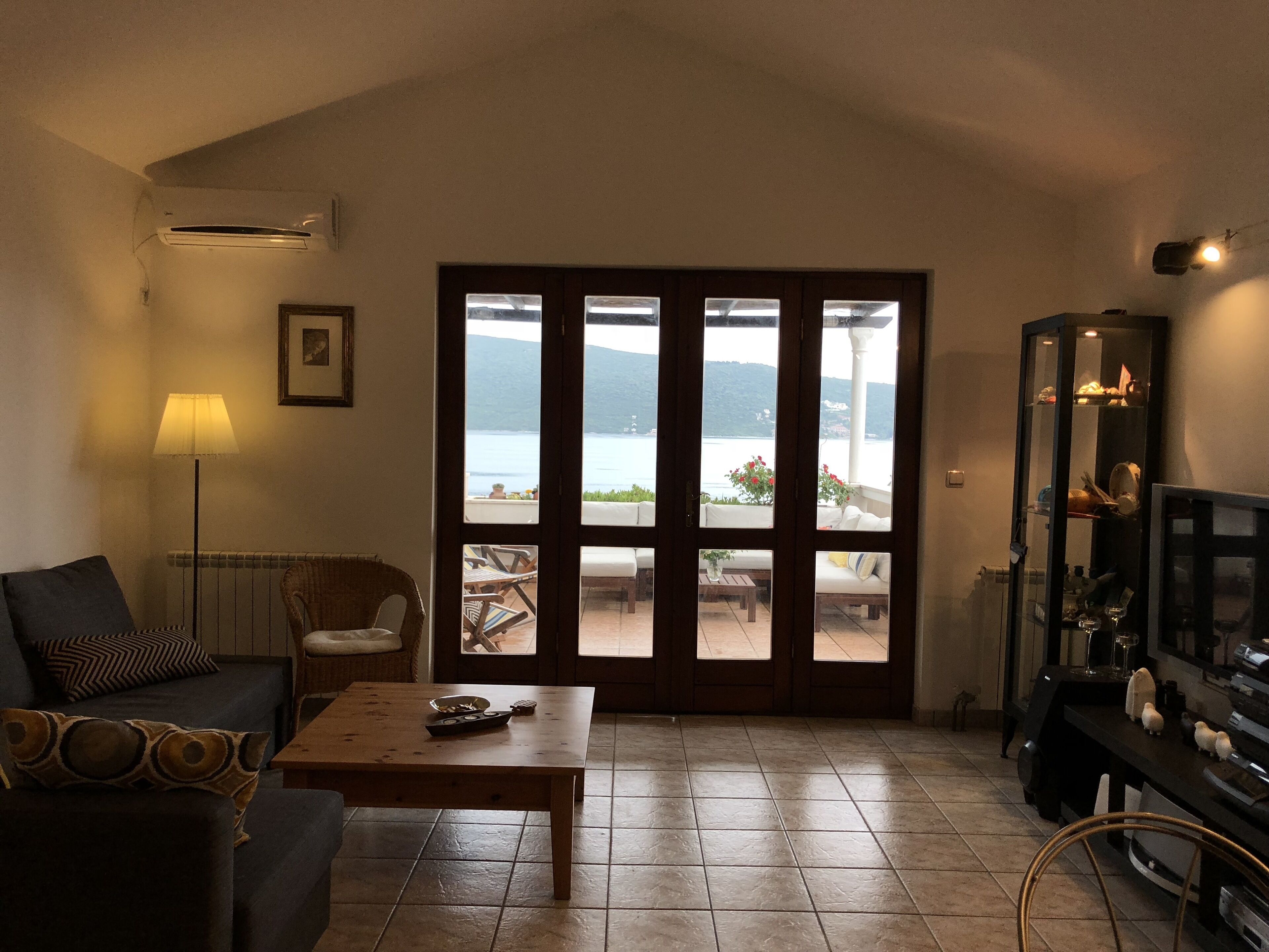 Ein Wohnzimmer mit einer Couch, einem Couchtisch und einer TV-Konsole. Ein großes Fenster bietet Blick auf das Meer und die Berge.