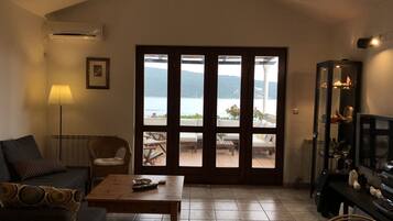 A living room with a sofa, a coffee table, and a TV stand. There is a large window with a view of the sea and mountains.