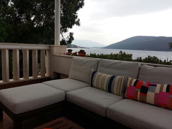 A balcony with a sofa and cushions, overlooking a body of water and hills.