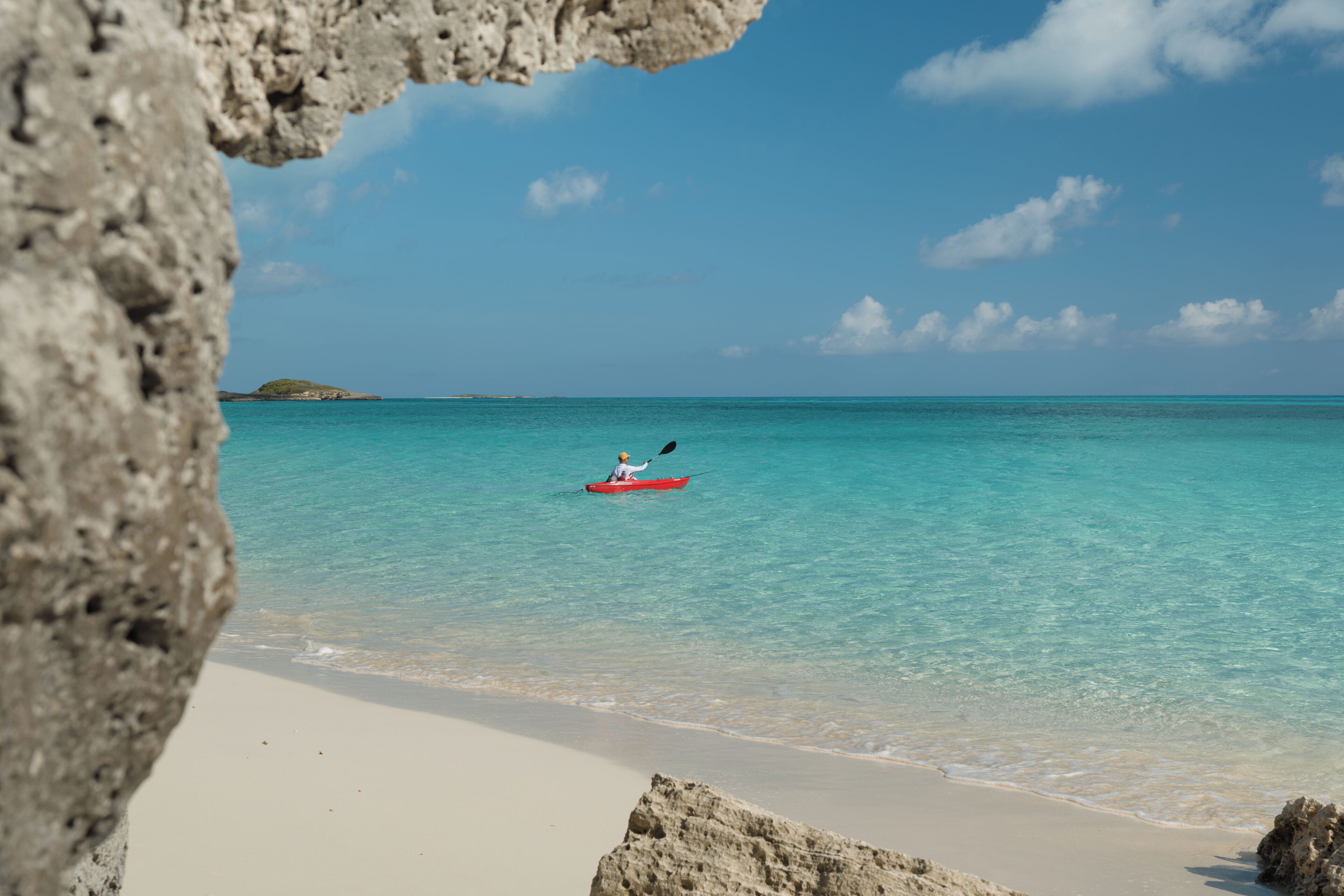 Ein Strand mit klarem türkisblauem Wasser, einer Person im Kajak und einem felsigen Vordergrund.