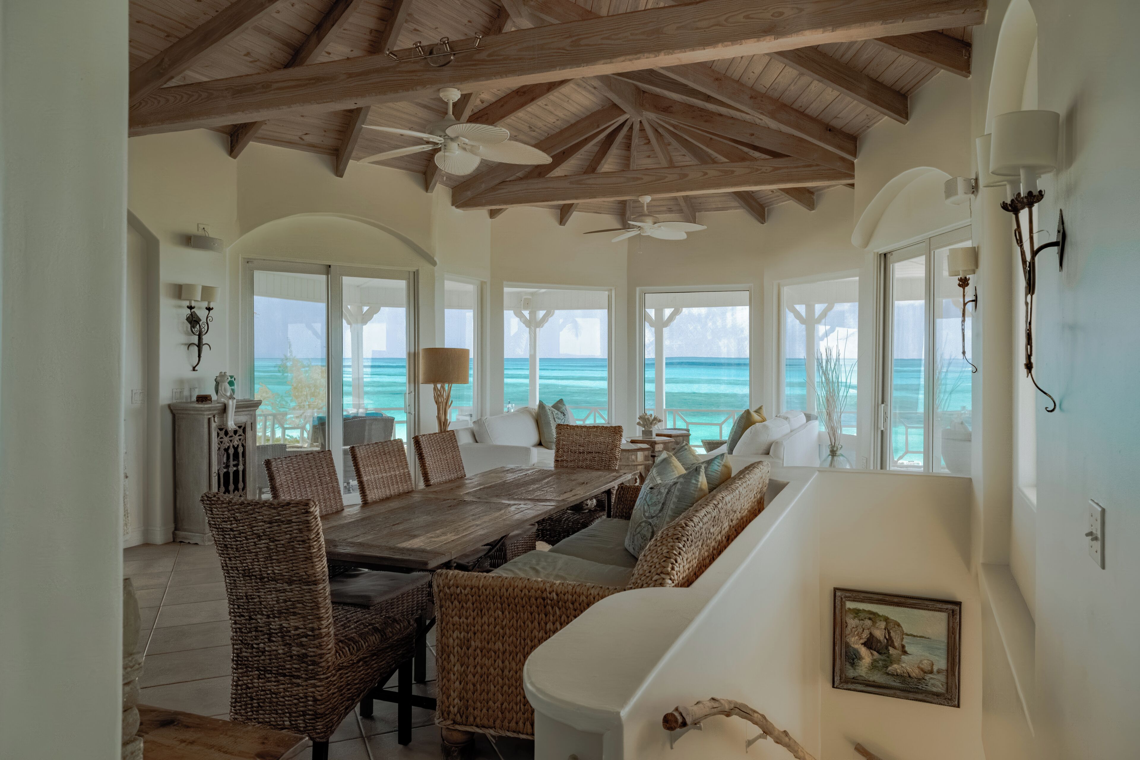 A spacious room with a wooden ceiling, large windows, a dining area with wicker furniture, and a view of the ocean.
