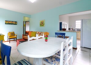 A dining area with a white table and blue chairs, a colorful sofa, and a kitchen in the background.