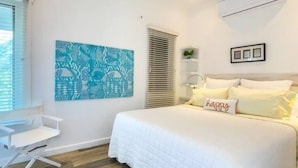 Un dormitorio con cama, una silla blanca, una ventana con persianas, una obra de arte en la pared y una mesita de noche.