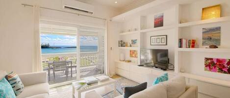 A spacious living room with a large window overlooking a beach, a fireplace, and a bookshelf filled with books and decorative items.