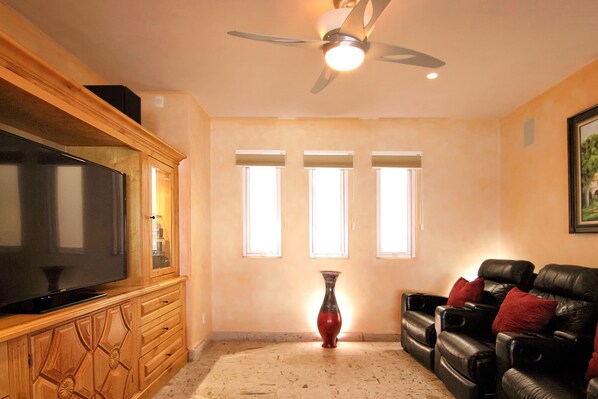 Una sala de estar con televisor de pantalla plana, ventilador de techo, chimenea y una zona de estar con sillones reclinables de cuero.