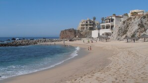 A sandy beach with a rocky shoreline, clear blue water, and buildings in the background. - Luxury Oceanfront Villa in Cabo San Lucas (Cabo San Lucas)