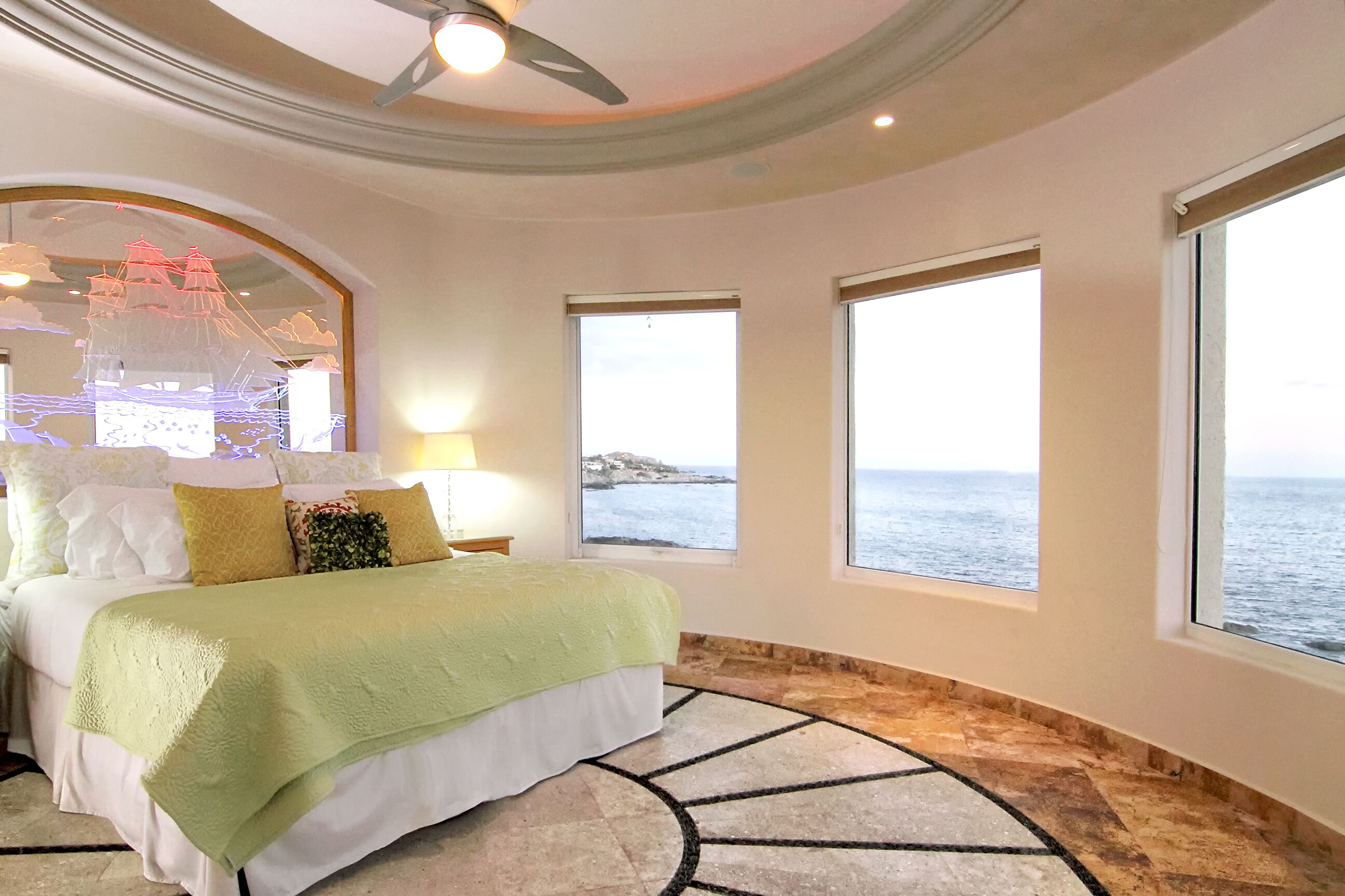 A bedroom with a bed, a nightstand, a lamp, a large mirror, and three windows with a view of the sea.