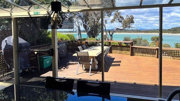 A sunlit room with a large glass window overlooking a wooden deck and a beach. - Beachfront Retreat with Direct Beach Access | Family Friendly (Greens Beach)