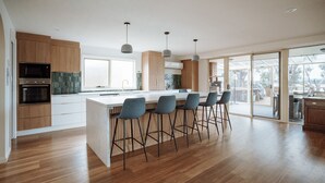 A modern kitchen with wooden cabinets, a central island with bar stools, and large windows. - Beachfront Retreat with Direct Beach Access | Family Friendly (Greens Beach)
