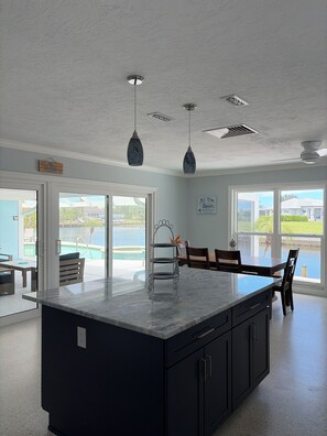 Une cuisine dotée d’un grand îlot central, de luminaires suspendus et offrant une vue sur une piscine et une plage.