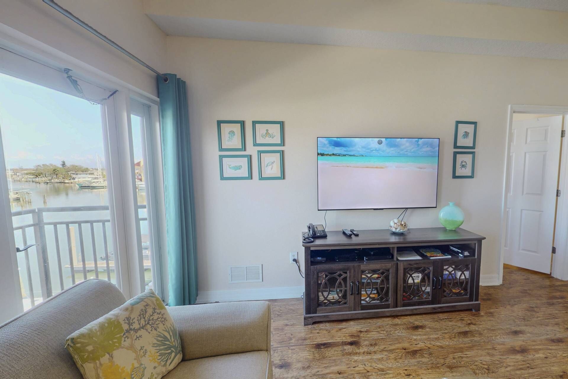 Ein Wohnzimmer mit großem Fernseher, gerahmten Bildern, einer Couch und Blick auf ein Gewässer durch eine Schiebetür.