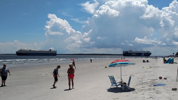 A beach with two large ships in the background, a clear sky, and people walking.