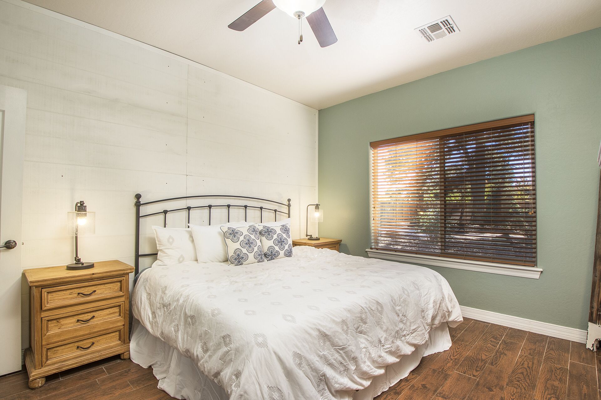 A bedroom with a bed, wooden nightstands, a ceiling fan, a window with blinds, and a wall with a textured pattern.