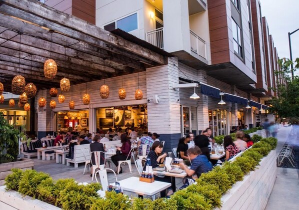 An outdoor dining area with tables and chairs, a restaurant entrance, and a building facade with a wooden overhang and hanging lights. - PCM Studio · Floor-to-Ceiling Views · Walk to BeltLine (Atlanta)
