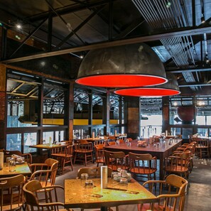 Un restaurante moderno con techos altos, vigas a la vista y amplios ventanales. Las mesas están dispuestas en un espacio amplio con sillas color naranja.