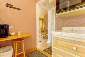 Una pequeña kitchenette con cafetera, un cubo de basura y un taburete de madera.