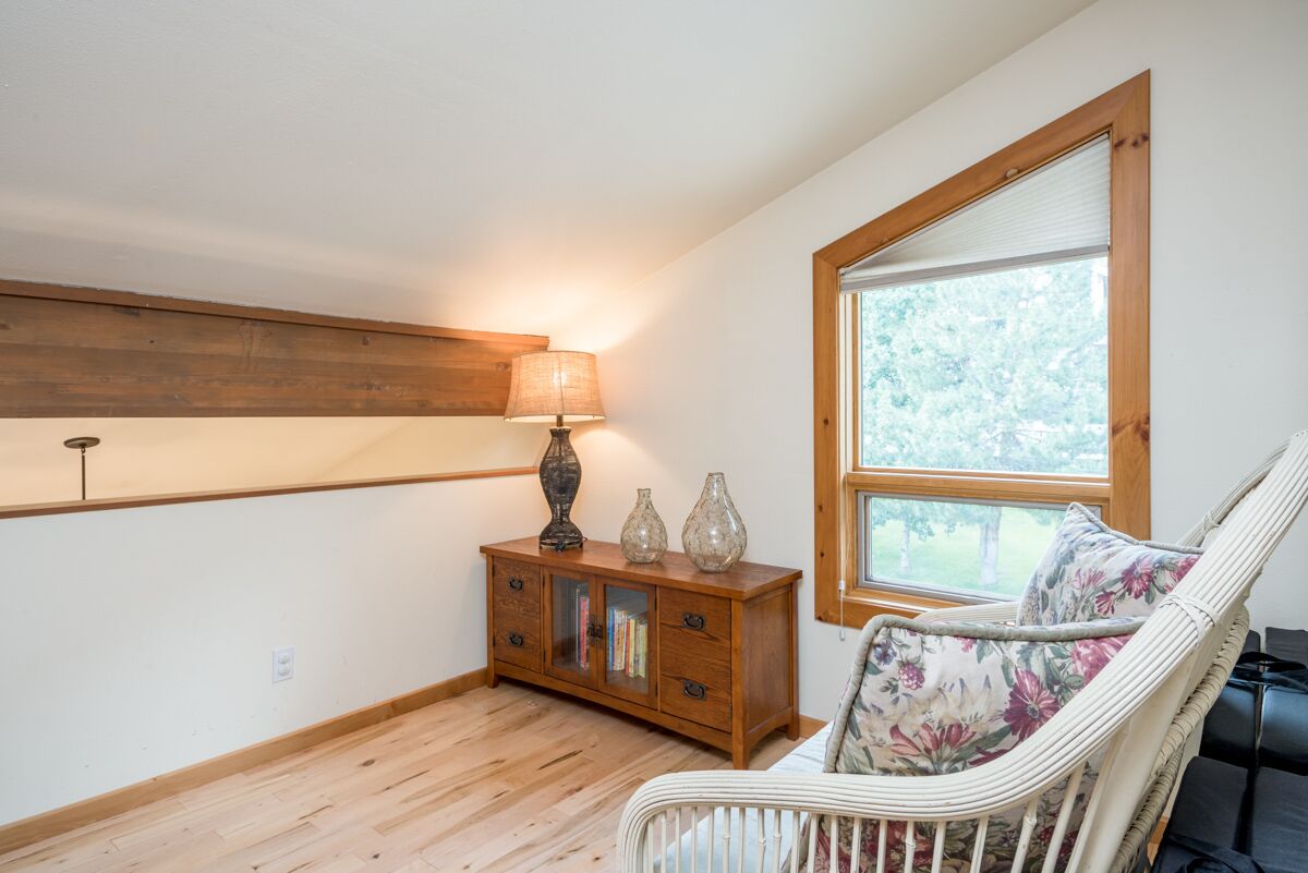 A room with a wooden floor, a white chair with floral cushions, a wooden cabinet with glass doors, and a window with a view of trees.