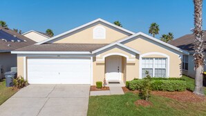 A single-story house with a white garage door, a small front yard with a lawn and a few shrubs, and a palm tree to the right.