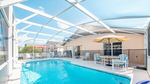 A covered swimming pool with lounge chairs and a table, surrounded by a glass enclosure.