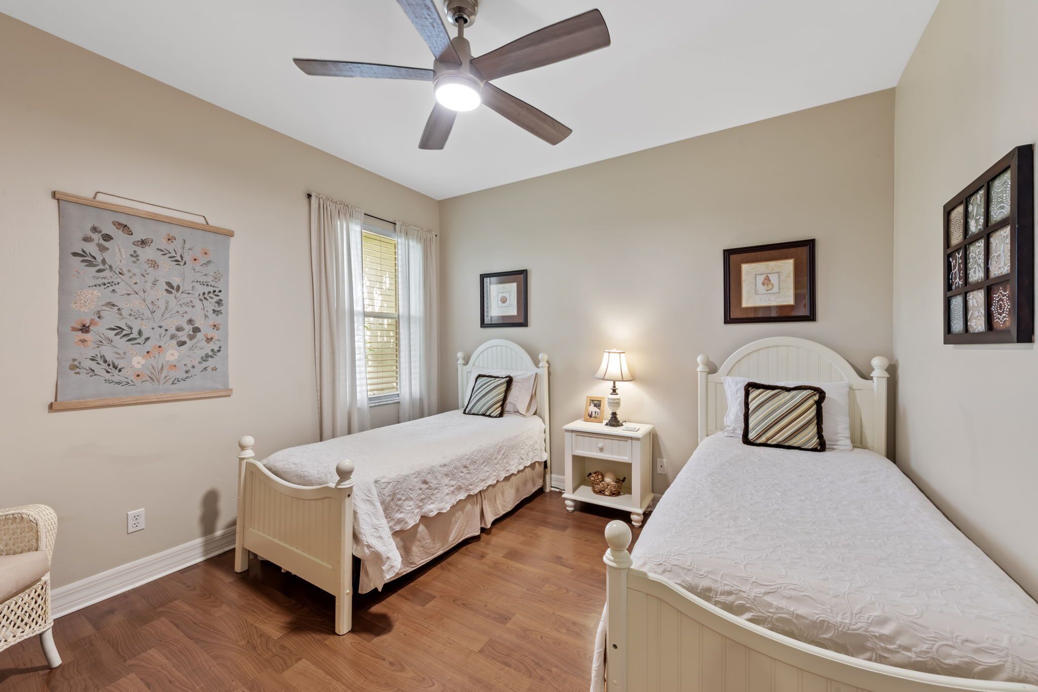 A bedroom with two single beds, a ceiling fan, a nightstand with a lamp, and a decorative wall hanging.