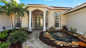 A house with a stone pathway leading to a front door, a small pond with rocks, and palm trees. - Villa Portofino: Resort Style Pool, Gulf Access (Cape Coral)