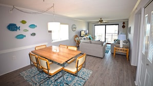 A dining area with a wooden table and chairs, a living room with a sofa and a TV, and a balcony with a view. - Cute, casual 2-bedroom condo with free WiFi and a partial ocean view located midtown and less than a block to the beach! (Ocean City)
