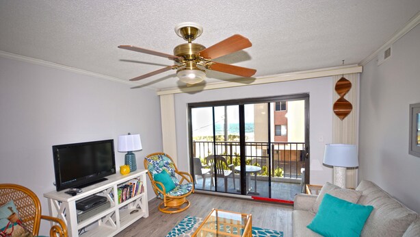 A living room with a television, bookshelf, wicker chair, sofa, and a ceiling fan. - Cute, casual 2-bedroom condo with free WiFi and a partial ocean view located midtown and less than a block to the beach! (Ocean City)