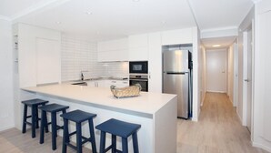 A modern kitchen with white cabinets, a stainless steel refrigerator, a microwave, and a large island with bar stools. - 2 Bedroom Apartment with pool and reverse Air conditioning (Mooloolaba)