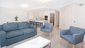 A modern living room with a grey sofa, two armchairs, and a white coffee table. A kitchen with white cabinets and a stainless steel refrigerator is visible in the background. - 2 Bedroom Apartment with pool and reverse Air conditioning (Mooloolaba)