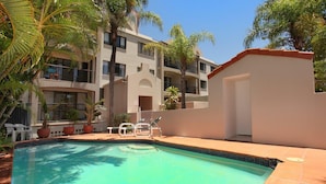 A pool area with a clear blue pool, surrounded by palm trees and a building with balconies. - 2 Bedroom Apartment with pool and reverse Air conditioning (Mooloolaba)