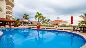 A swimming pool with lounge chairs and umbrellas, surrounded by palm trees and a building with a dome. - Bienvenido Amigo! In the heart of Amapas  2BD Condo for rent in Old Town, Puerto (Puerto Vallarta)