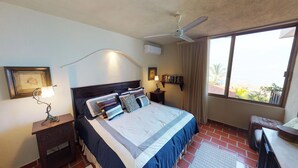 A hotel room with a bed, a nightstand, a desk, a ceiling fan, a window with a view, and a wall-mounted air conditioner. - Bienvenido Amigo! In the heart of Amapas  2BD Condo for rent in Old Town, Puerto (Puerto Vallarta)