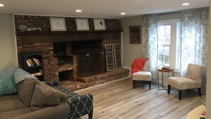 A cozy living room with a brick fireplace, a bookshelf, two armchairs, and a window with patterned curtains.