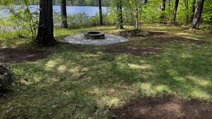 A campsite with a fire pit surrounded by gravel, trees, and a lake in the background. - Waterfront Chalet with private beach in the White Mountains of New Hampshire. (Conway)