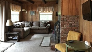 A cozy living room with a wooden ceiling, a brick fireplace, a sectional sofa, a small table, and a television mounted on the wall. - Waterfront Chalet with private beach in the White Mountains of New Hampshire. (Conway)