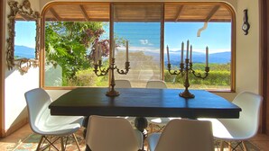 Un comedor con una mesa y sillas, vistas a zonas verdes y montañas a través de amplios ventanales, y techo de madera.