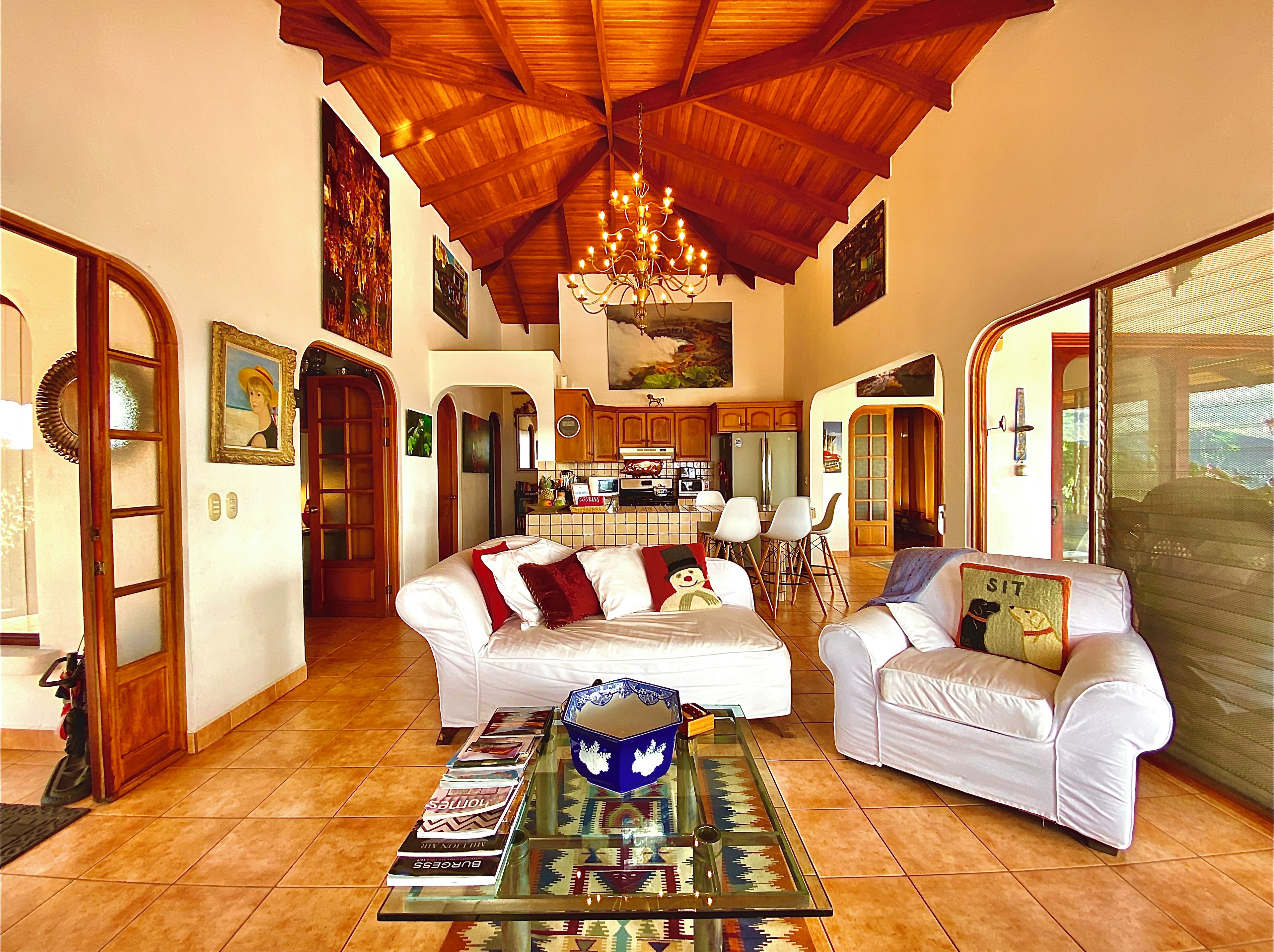 A spacious living area with a high wooden ceiling, a dining area with a kitchen in the background, and a large window with a view.