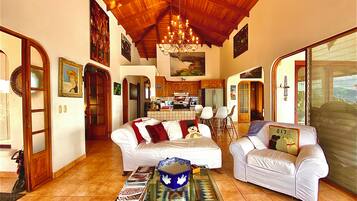 A spacious living area with a high wooden ceiling, a dining area with a kitchen in the background, and a large window with a view.