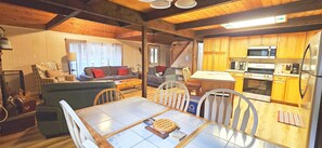 Interior acolhedor de uma cabana com mesa de jantar, cadeiras, sofá, cozinha equipada e escada.