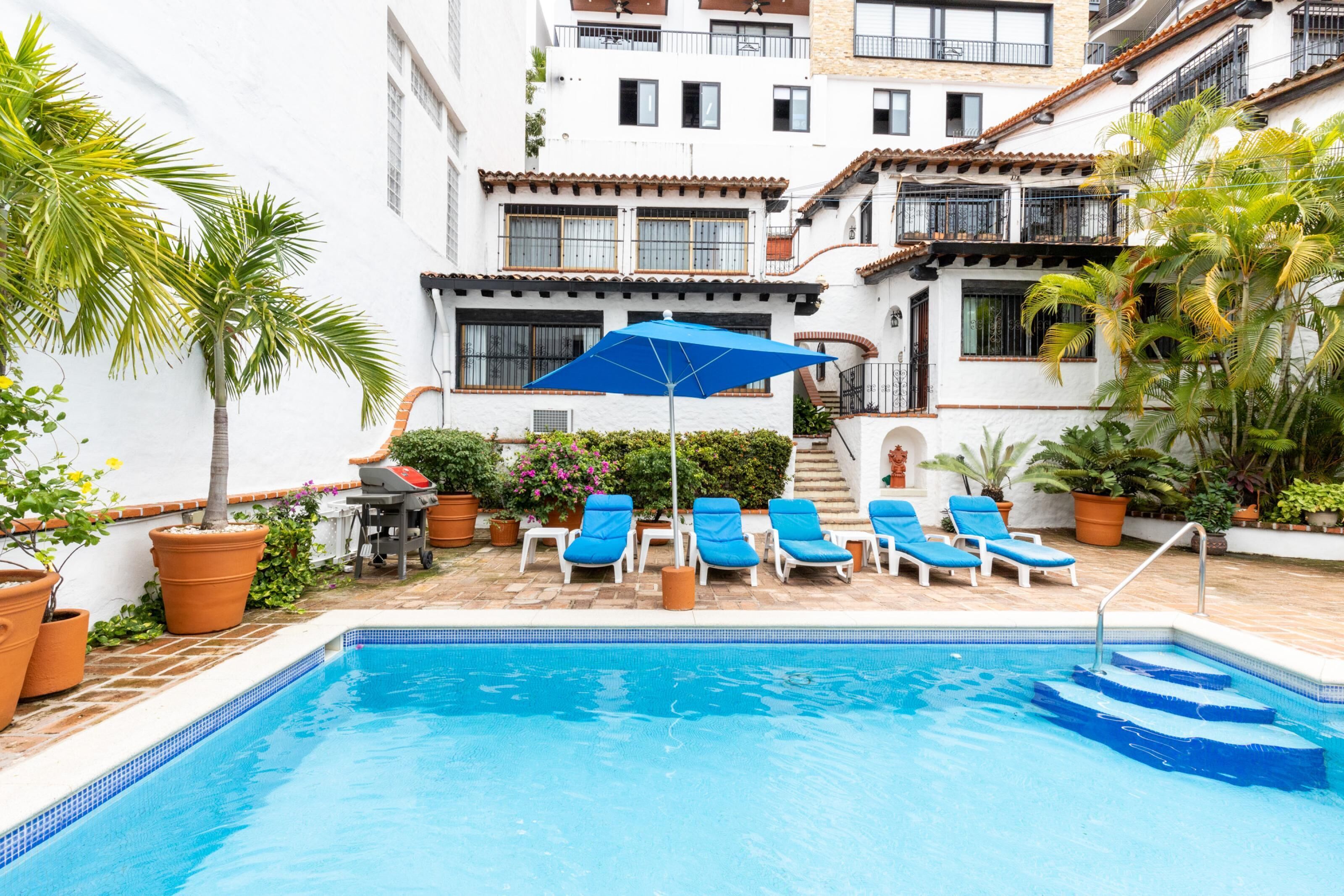 Una piscina con sillas reclinables azules y una sombrilla, rodeada de plantas en macetas y un edificio blanco con balcones.