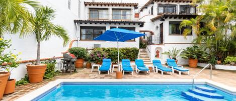 Una piscina con sillas reclinables azules y una sombrilla, rodeada de plantas en macetas y un edificio blanco con balcones.