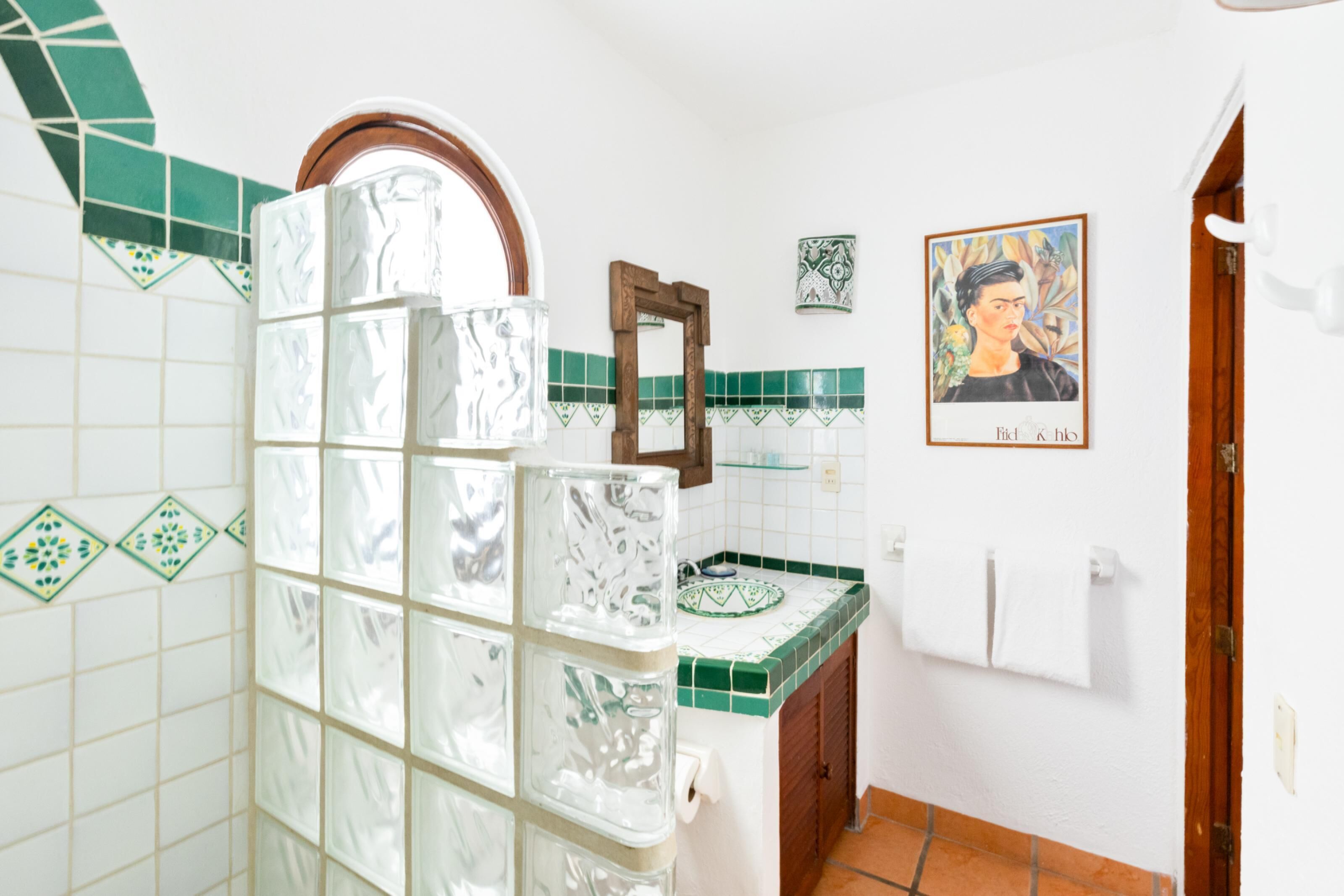 A bathroom with white and green tiled walls, a glass shower enclosure, a wooden door, and a framed artwork on the wall.