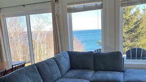 Un salon doté d’un canapé d’angle gris, de grandes fenêtres équipées de stores, d’un tapis à motifs et d’une vue sur la mer.
